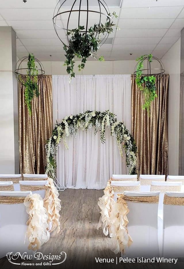 A Gold And white backdrop sits behind a white moongate arch decorated in draping florals of green and white. With hanging iron Orbs draped in greenery extending from the ceiling. And an aisle of fitted white spandex covered chairs, with a gold sequined band. The inner chairs adorned with a hanging tuft of nude material swirling to the ground.